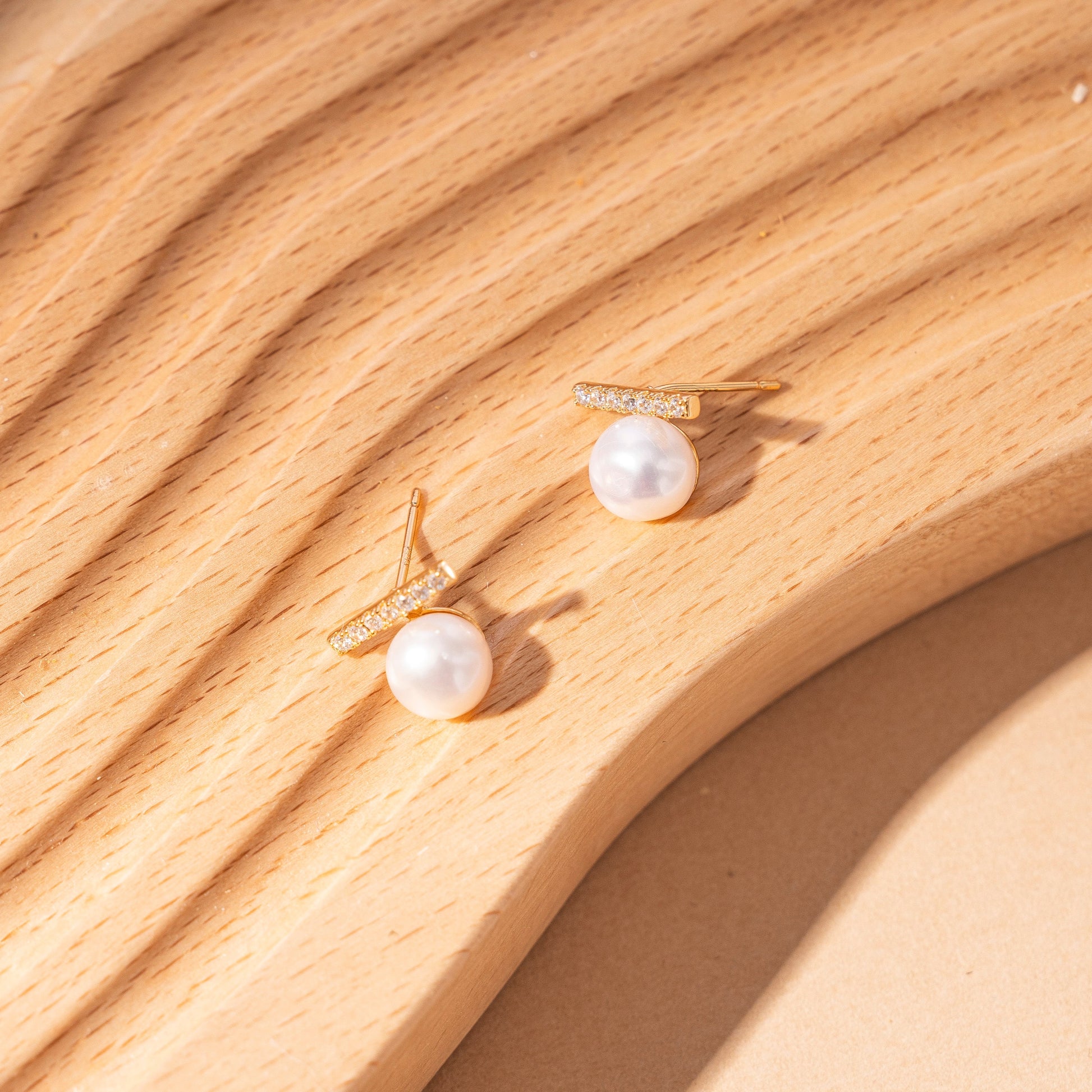 STELLA MARGARITA balance beam pearl stud earrings with gold and crystal detailing on wooden surface
